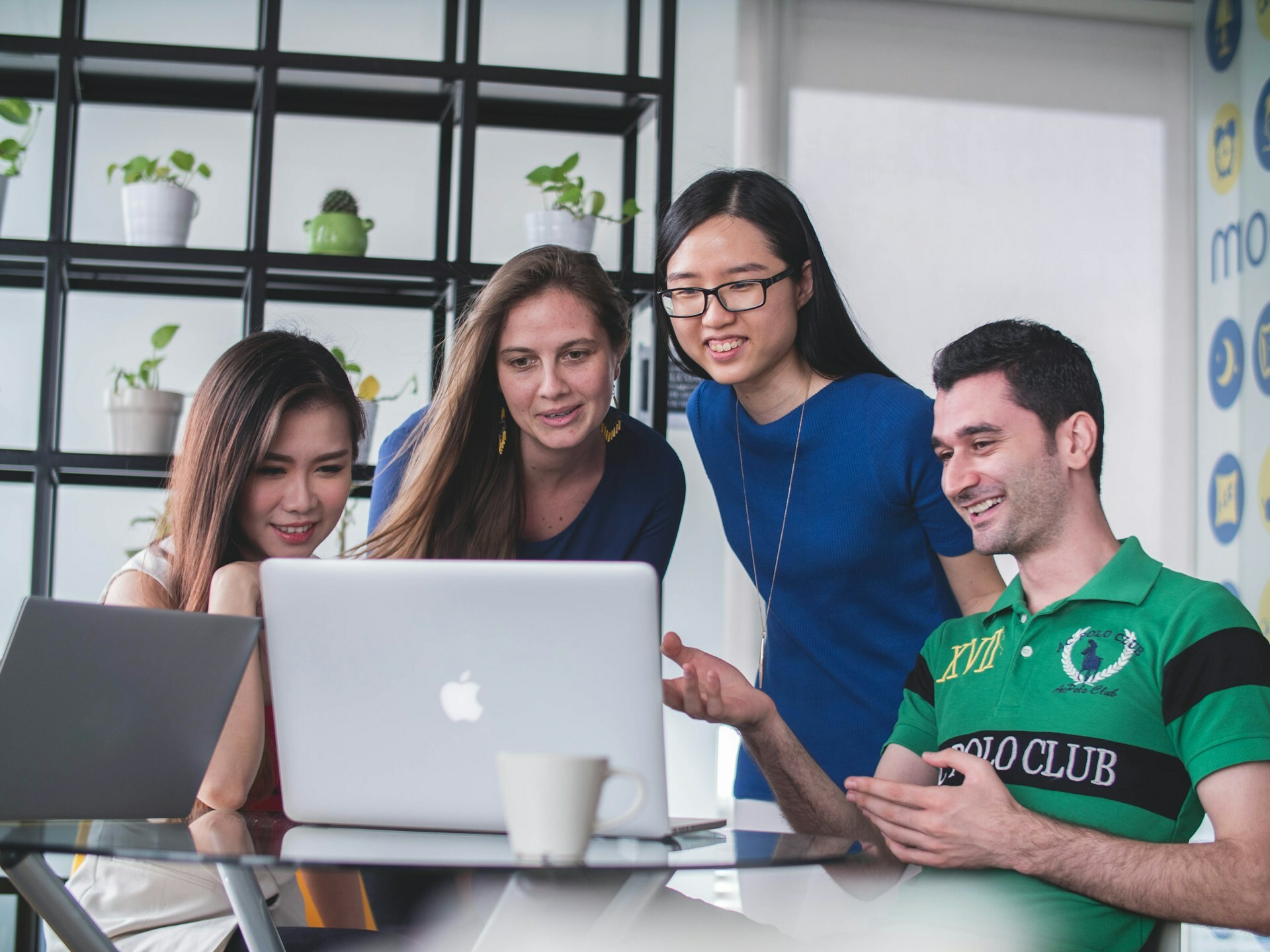 A group of young people are sitting around a laptop and looking at the screen in an office environment.