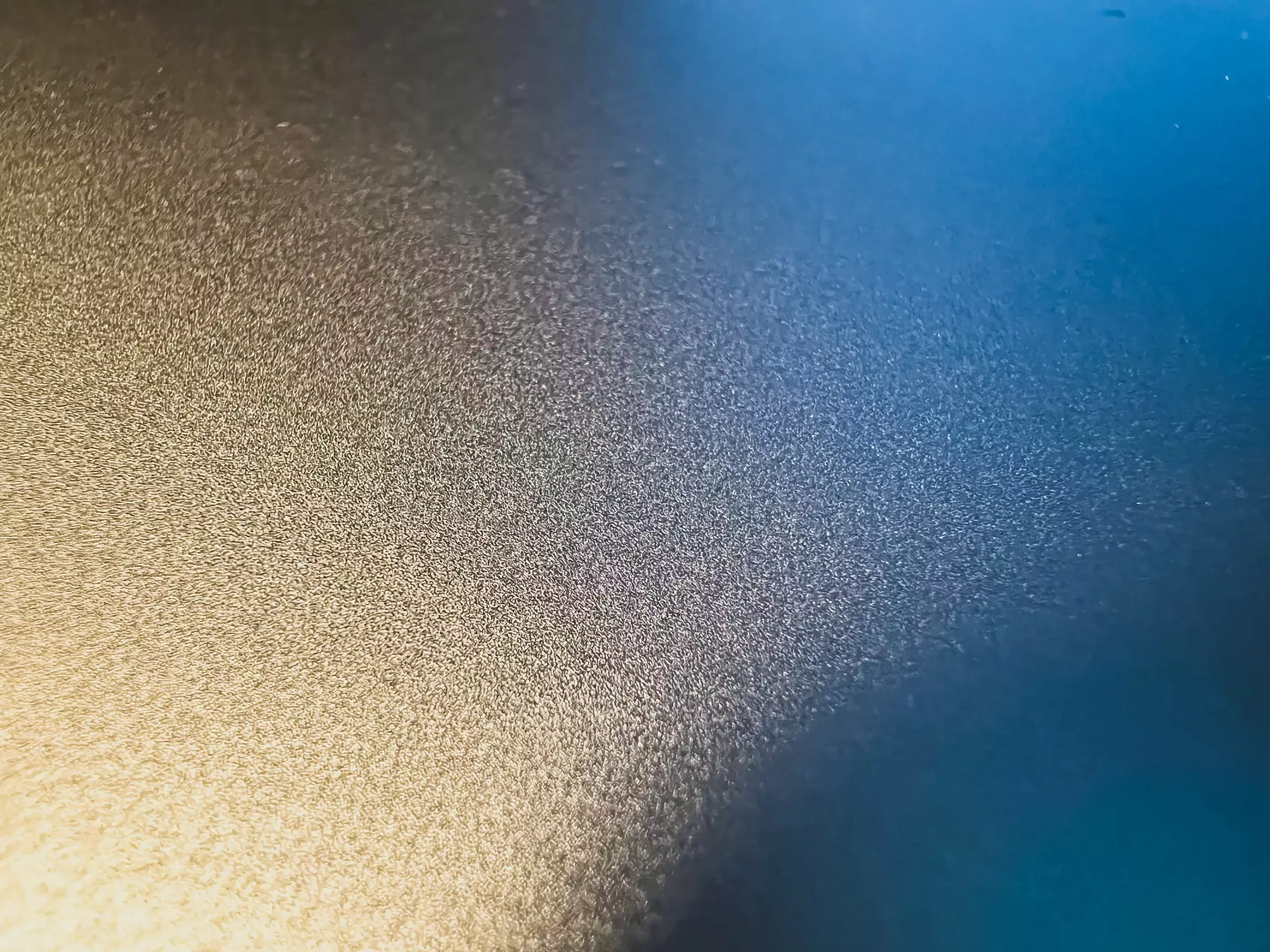 Close-up of a metallic surface with a subtle gradient from golden on the left to blue on the right. The texture appears rough, creating a shimmering effect.